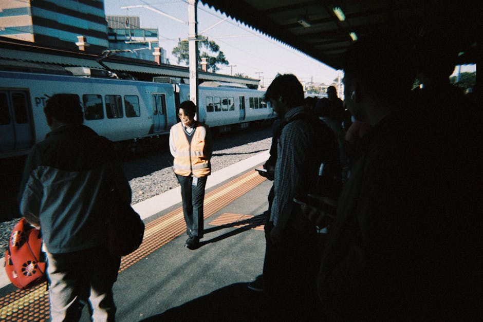 At a train station platform near Blackheath Station, several commuters are waiting for a train. The scene shows a woman wearing a high-visibility vest standing near the edge of the platform, with her