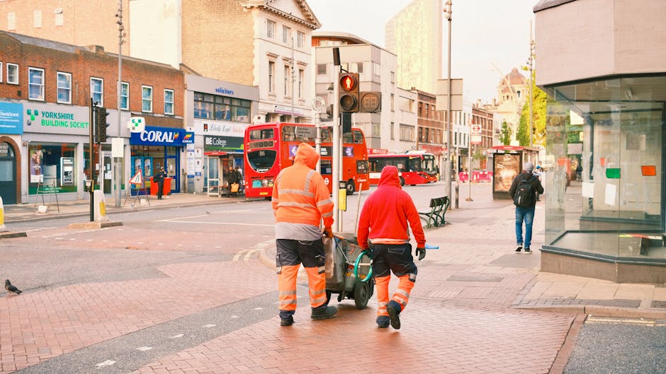 At a train station platform near Blackheath Station, several commuters are waiting for a train. The scene shows a woman wearing a high-visibility vest standing near the edge of the platform, with her